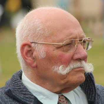 Old and bald man with some gray hair and large moushtache. He is wearing golden frame eye glasses and looks into distance. With serious face.